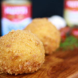 two arancini balls on a wooden platter with sauce and tomatoes in the background